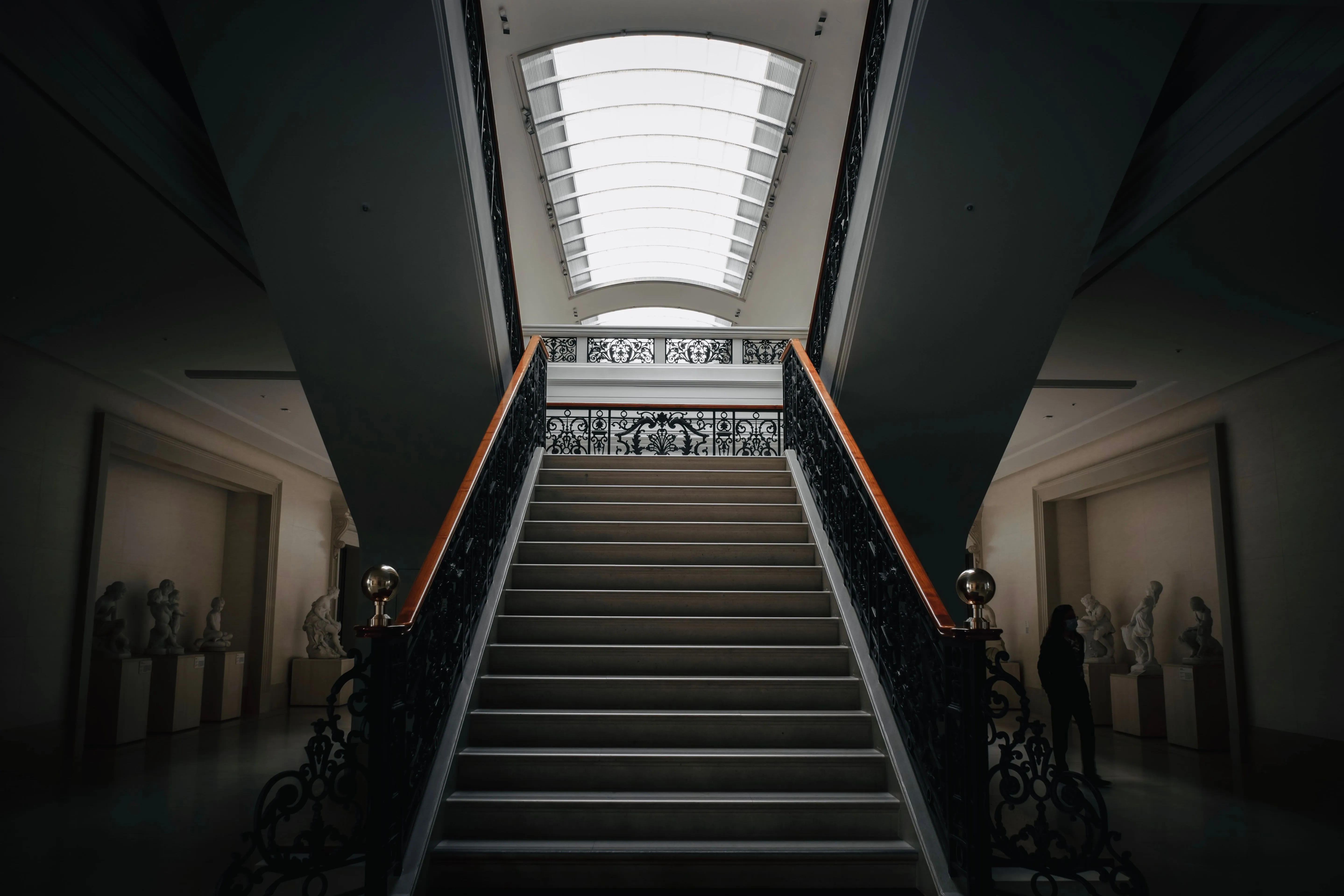 A dim museum stair hall under a skylight, sculptures standing in alcoves on either side