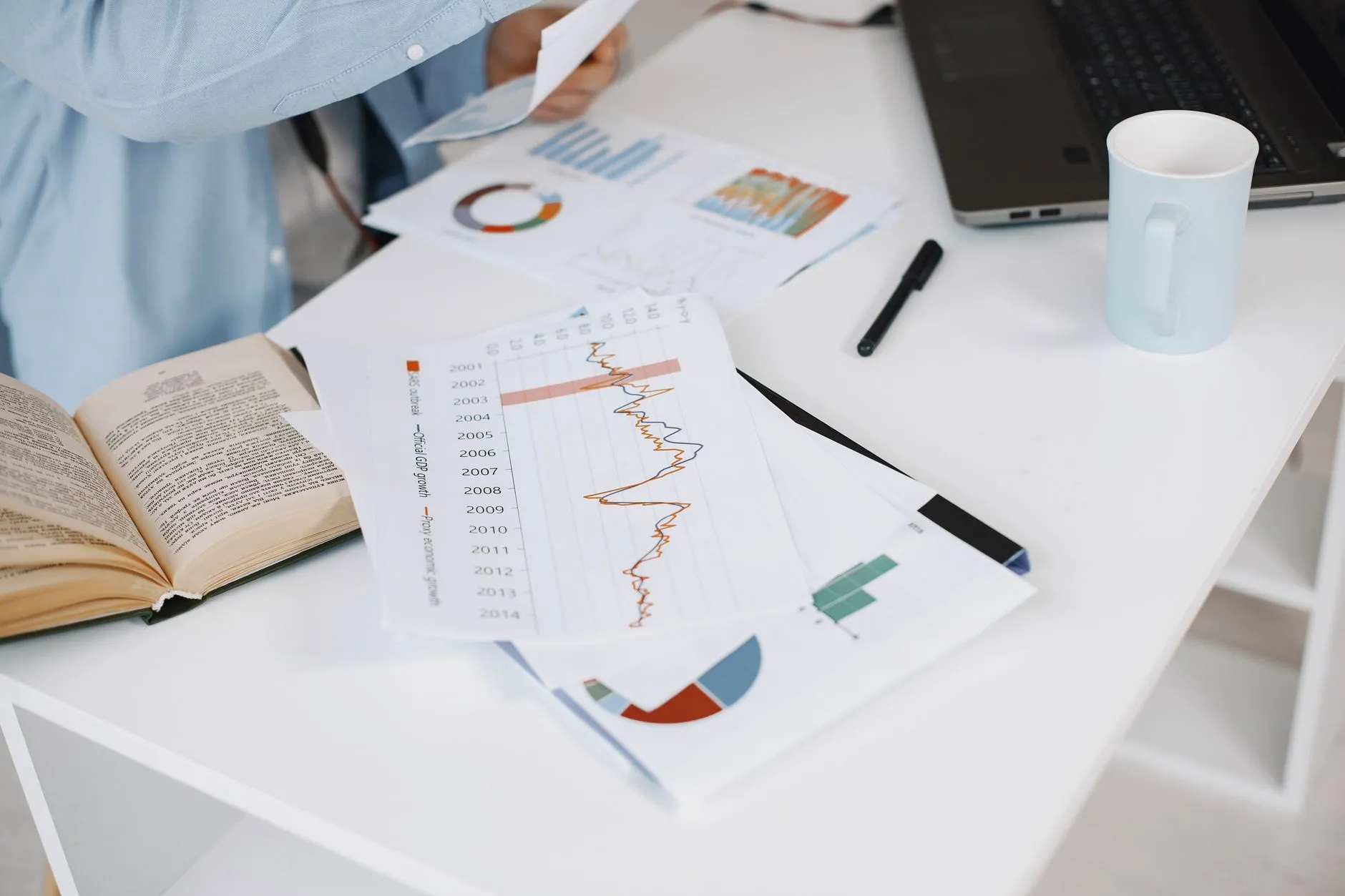 Printed charts, an open book, and a laptop on a white desk. Photo by Gustavo Fring / Pexels.